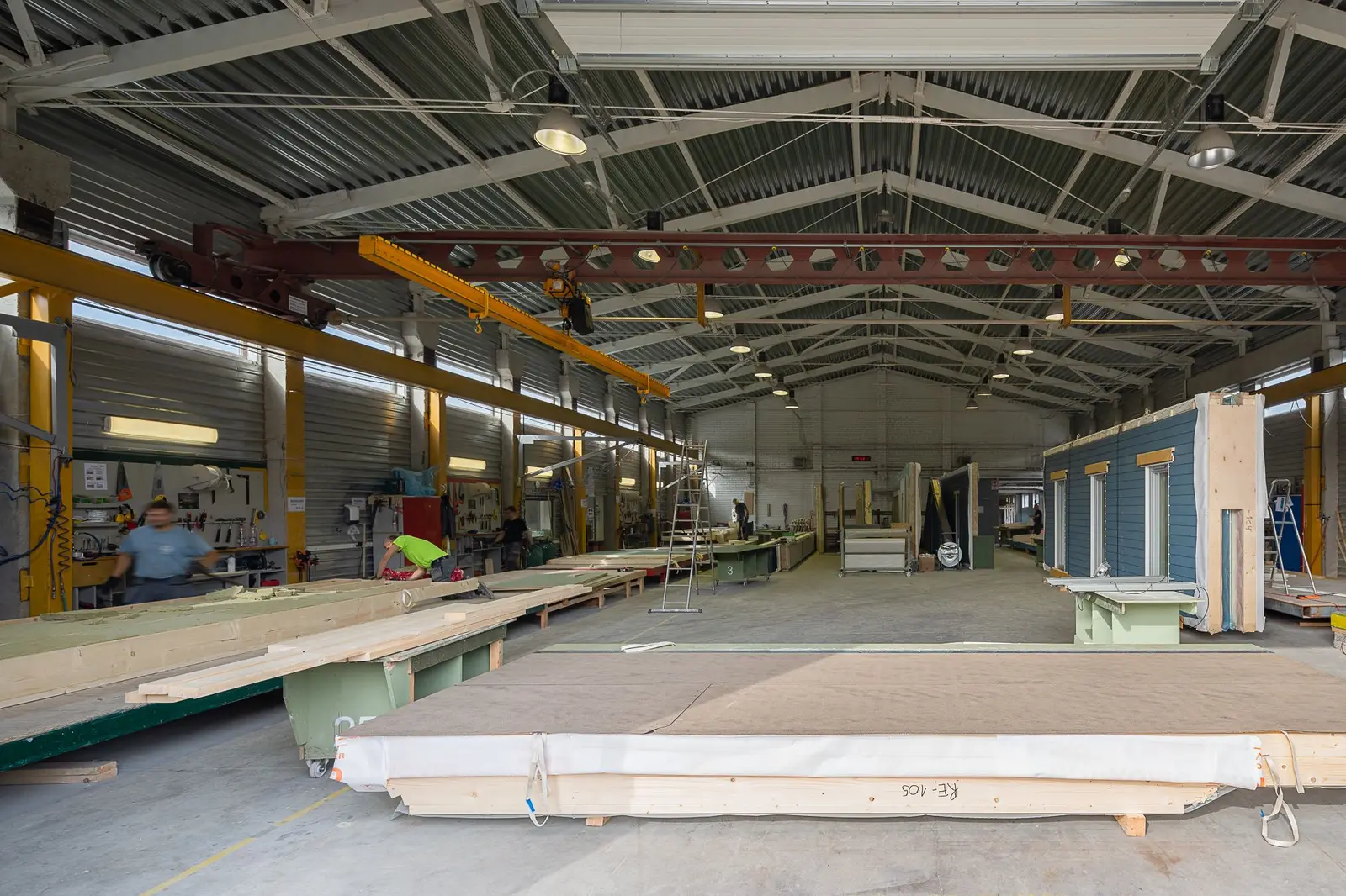 Inside the element factory: timber wall and floor elements on assembly lines beneath an overhead gantry crane, with a finished module wall standing at the right