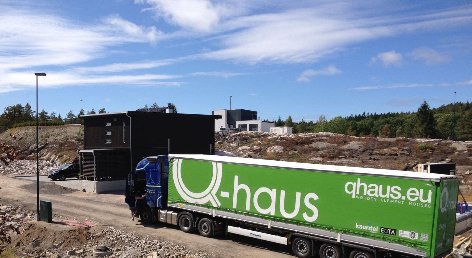 A curtain-side truck delivers prefabricated house elements to a rocky building site, the first house already standing behind it